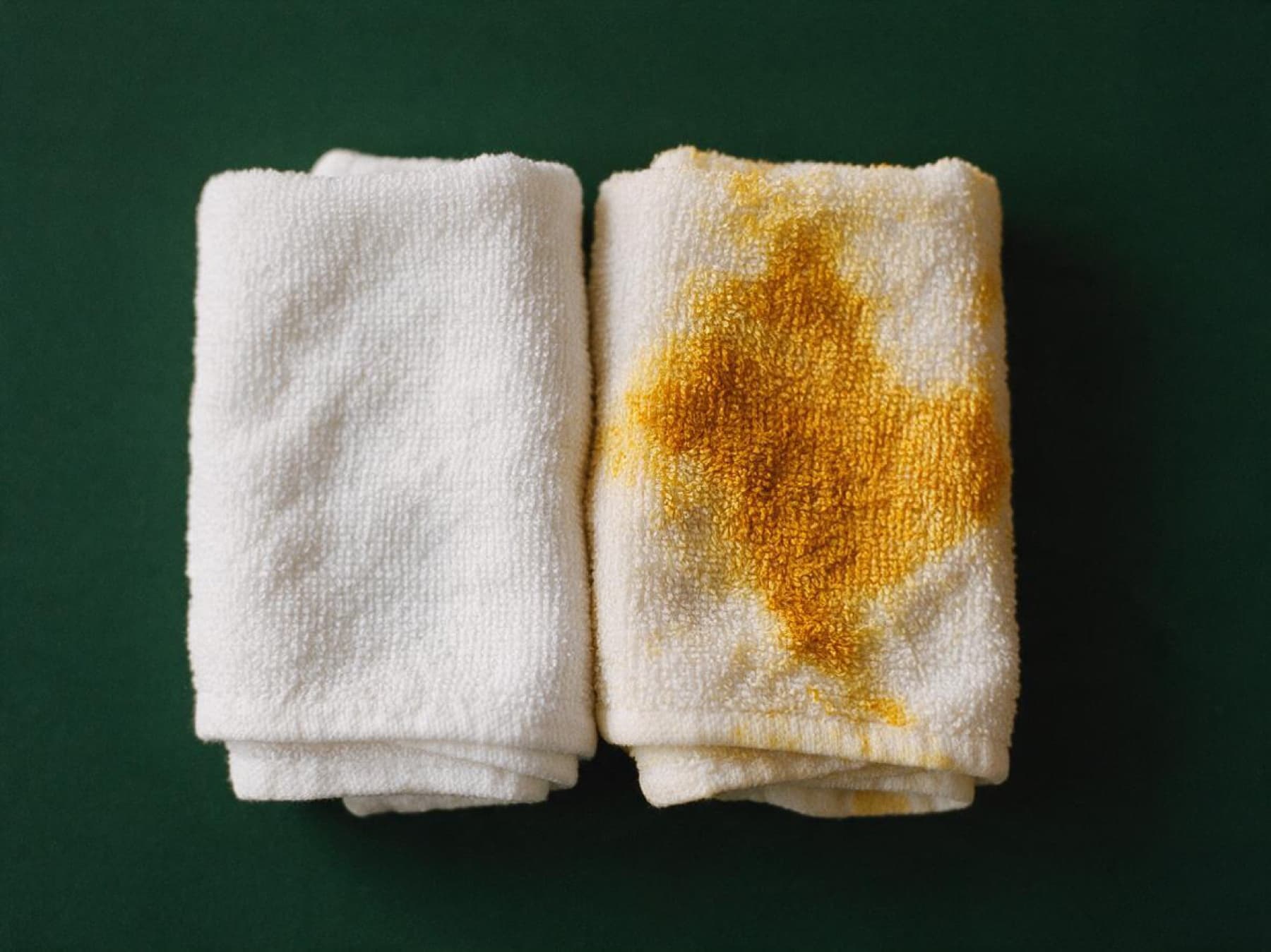 Two folded white washcloths on a dark green surface. The one on the right is stained golden by turmeric.